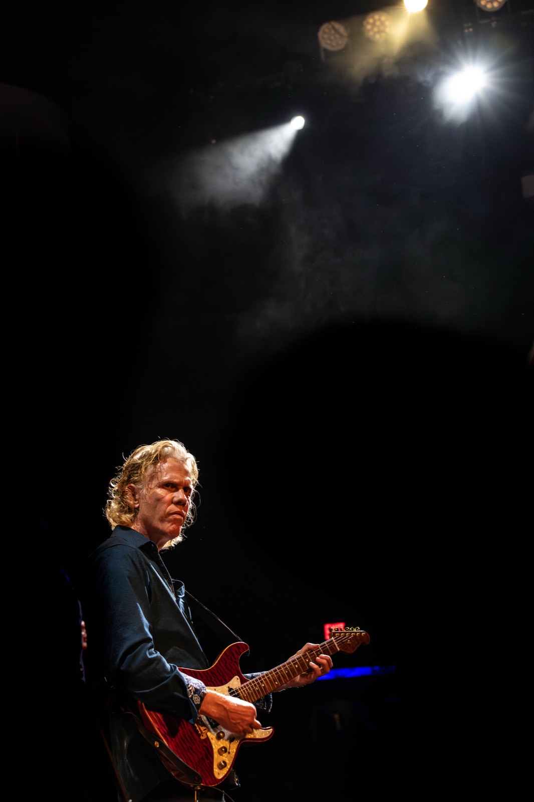Erik Madsen standing alone on stage under two dramatic spotlight beams, vast darkness above