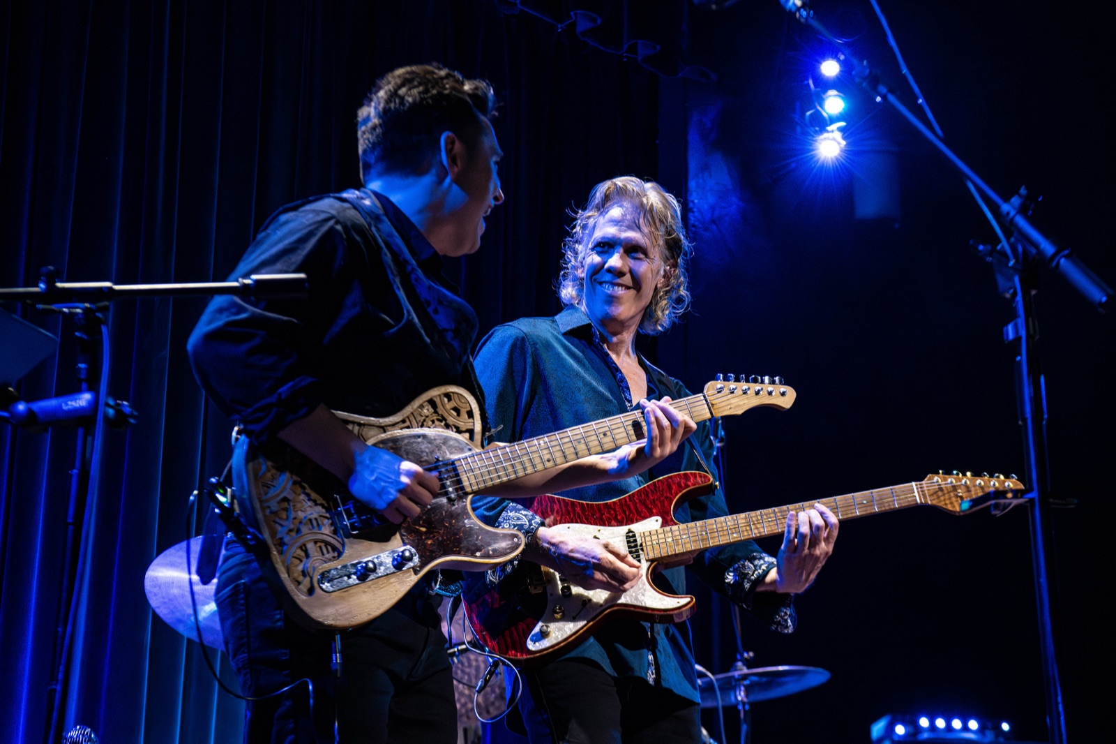 Two-guitar moment on stage under electric blue lighting, Erik beaming